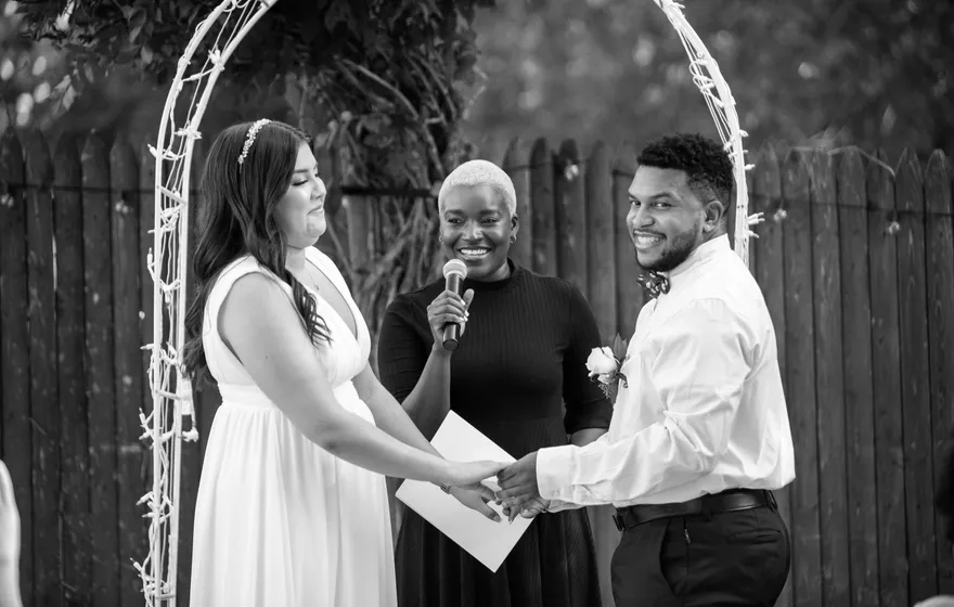 Black and white photo of an officiant holding a microphone and a sheet of paper while a bride and groom hold hands under a white arch