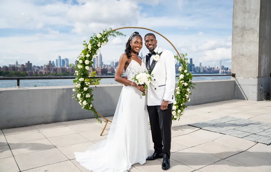 A bride and groom stand inside a round floral arch on a Brooklyn rooftop terrace, the Manhattan skyline across the river behind them