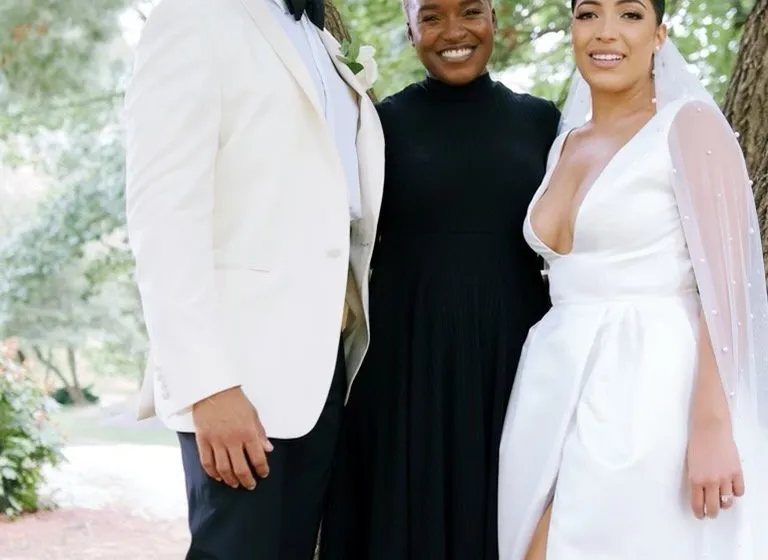 Robyn stands between a groom in a white dinner jacket and a bride in a satin gown under the trees near Cop Cot in Central Park