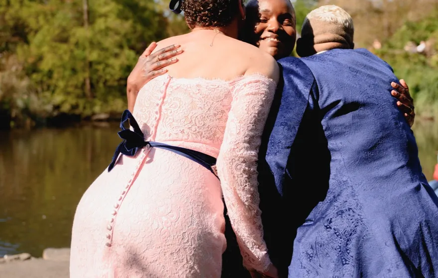 Two spouses embrace their officiant in a three-way hug beside the Pond in Central Park at a vow renewal