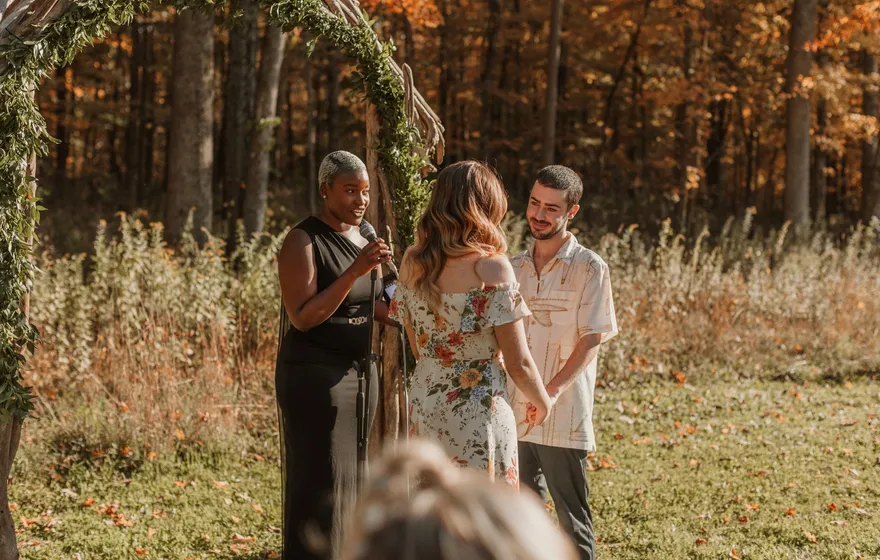 An officiant speaks at a microphone beside a couple standing under a branch arch in an autumn field