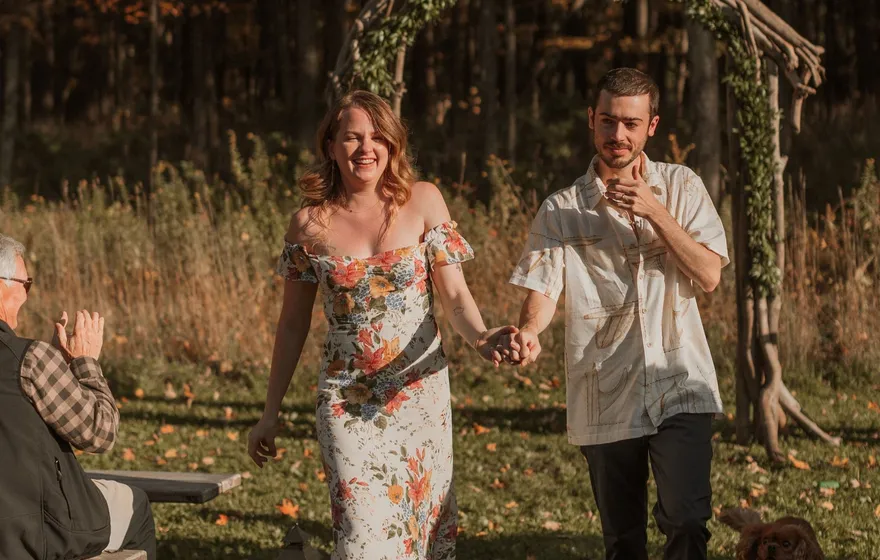 A laughing bride in a floral dress and a groom in a patterned shirt walk hand in hand away from a branch arch as a guest claps