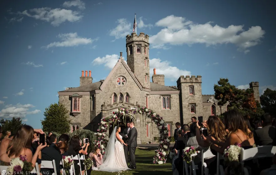 A couple holds hands under a tall flower arch on the lawn in front of Whitby Castle's stone tower, guests seated in rows on both sides of the aisle