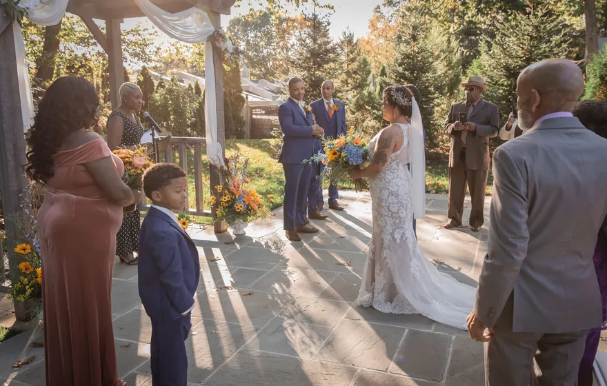 A bride in a lace gown holding a bouquet of sunflowers and roses reaches the front of an outdoor ceremony, where two men in blue suits wait and family members stand close by