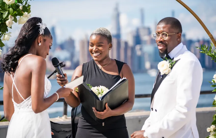 An officiant laughs while reading from a black folder as a couple exchanges vows on a rooftop, the Manhattan skyline and East River behind them