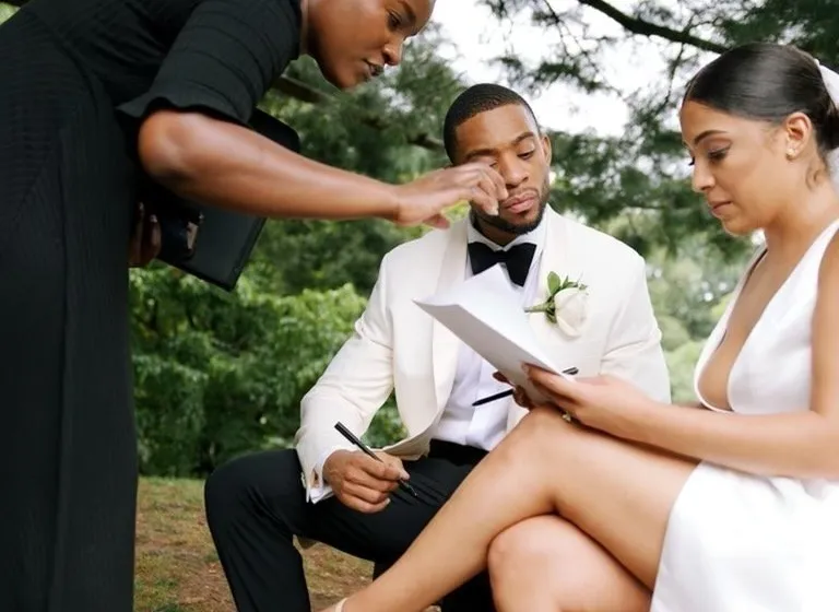 A seated couple holds pens and papers on a park bench while the officiant leans in toward them