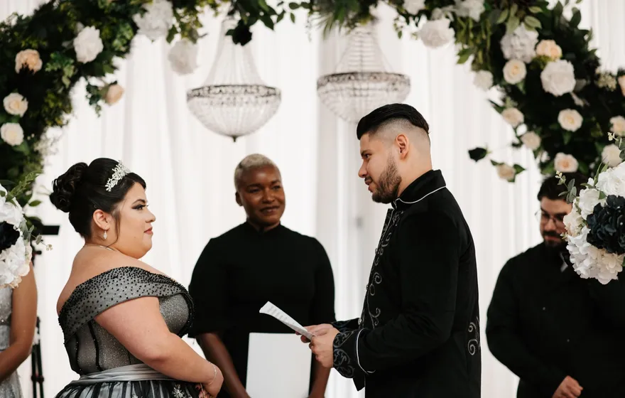 A groom reads his vows from a small card while the bride listens and the officiant stands between them under a floral arch