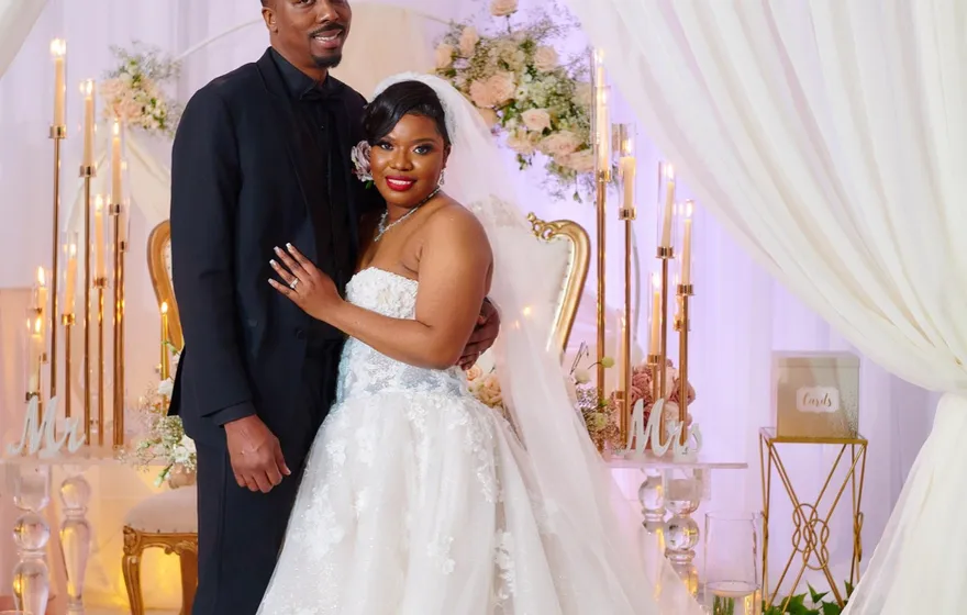 A bride in a full white gown and a groom in a dark suit stand in a Brooklyn loft in front of white drapes, flowers and rows of candles