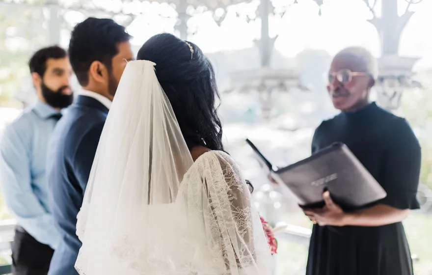 The officiant reads from a black binder to a bride in a long veil and a groom inside the Ladies Pavilion, a friend standing beside them
