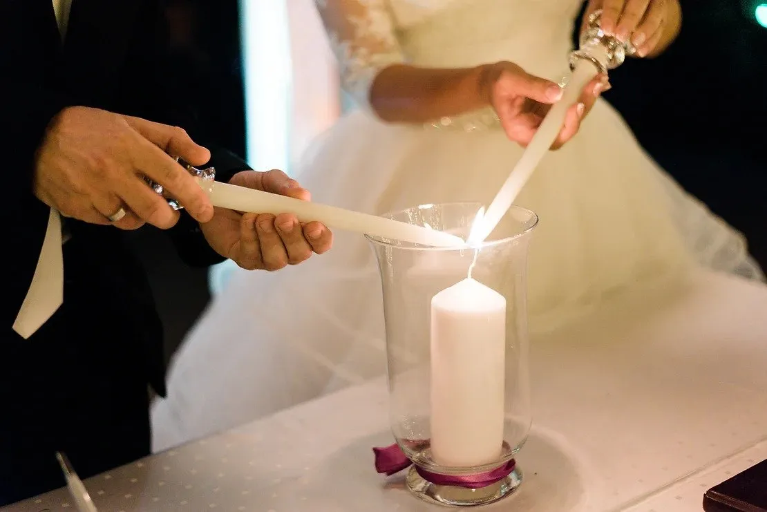 A bride and groom lowering two tapers into a glass hurricane vase to light a white pillar candle