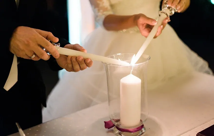 A bride and groom lower two tapers into a tall glass hurricane vase to light a white pillar candle protected inside, a magenta ribbon tied around the base