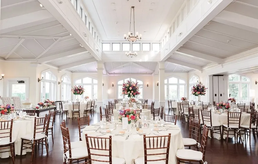 Whitby Castle's white ballroom set for a reception, with round tables, dark wood chairs, tall floral centerpieces and a row of arched windows