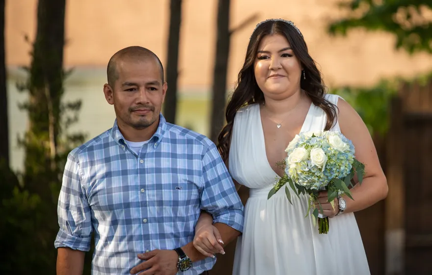 A bride holding a bouquet of white and blue hydrangeas walks on the arm of a man in a plaid shirt down a backyard aisle
