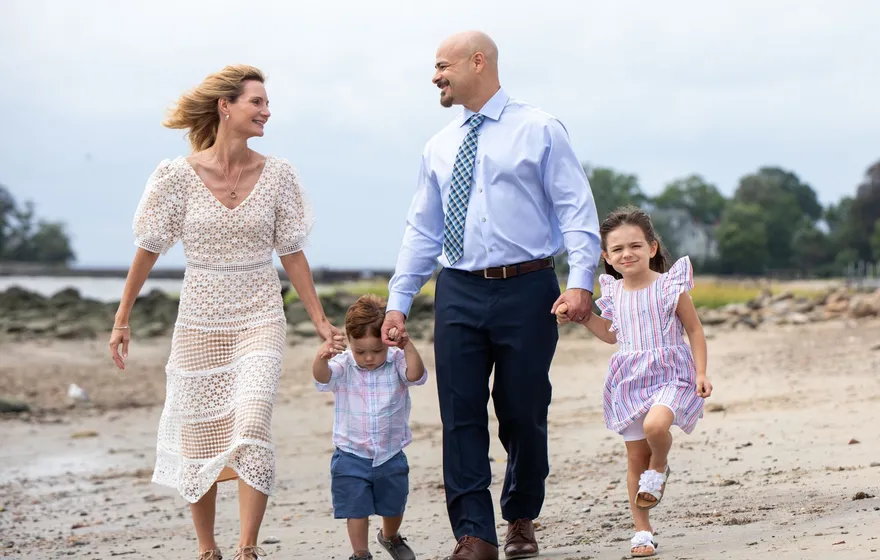 A couple walk hand in hand along a beach with a toddler boy and a young girl in a ruffled dress between them