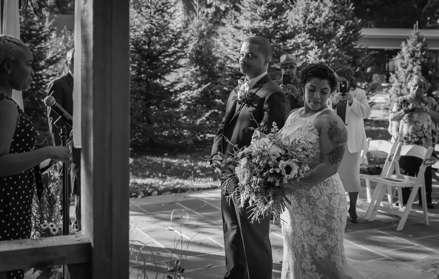 Black and white photo of a bride holding a large bouquet as she walks up a garden aisle on a man's arm, guests turned to watch