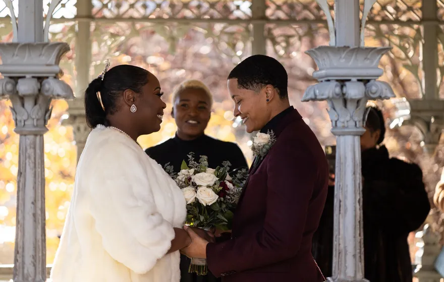 A bride in a white fur wrap and a partner in a burgundy suit face each other under the cast-iron roof of the Ladies' Pavilion while the officiant stands between them