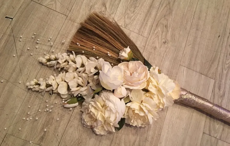 A wedding broom lying on a wood floor, the handle wrapped in metallic ribbon and the base dressed with cream roses, hydrangea, and scattered pearls