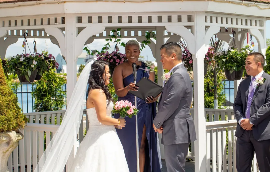 Under a white gazebo, the officiant reads from an open black folder into a microphone while the bride and groom face each other and a groomsman looks on