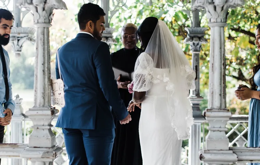 A bride in a long veil and a groom stand before the officiant inside the white cast-iron Ladies Pavilion in Central Park, a few guests around them