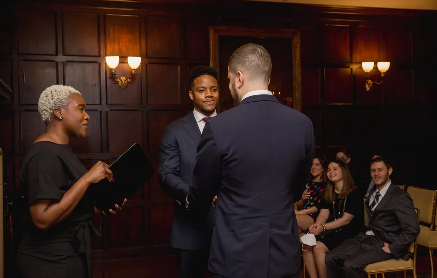 An officiant reads from a black binder to two grooms in suits in a wood-paneled room while a few guests watch from their seats