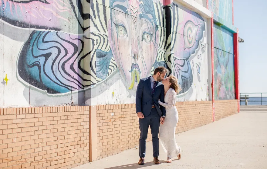 A bride and groom kiss in front of a large painted mural on a boardwalk