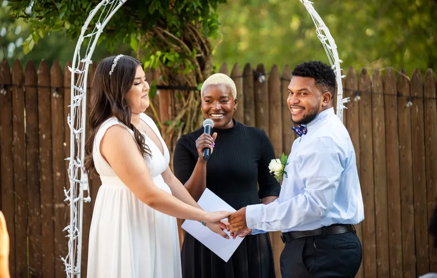 An officiant with a microphone and a printed script stands under a white wire arch between a bride and groom holding hands in a backyard