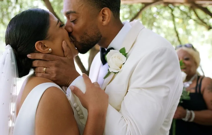 A bride and groom kiss under the rustic wooden roof of Cop Cot, a guest smiling just behind them