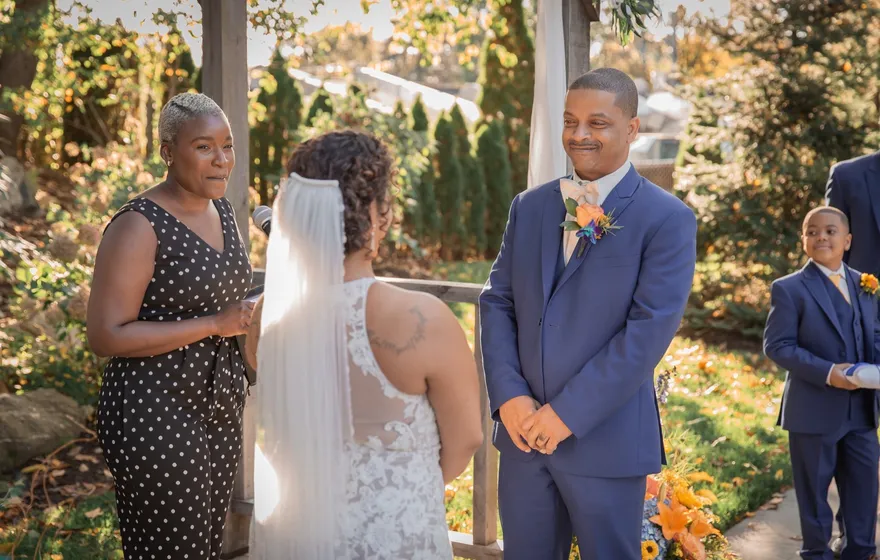 An officiant with short gray hair looks up from her notes and speaks to a smiling groom while the bride stands between them under a wooden arbor