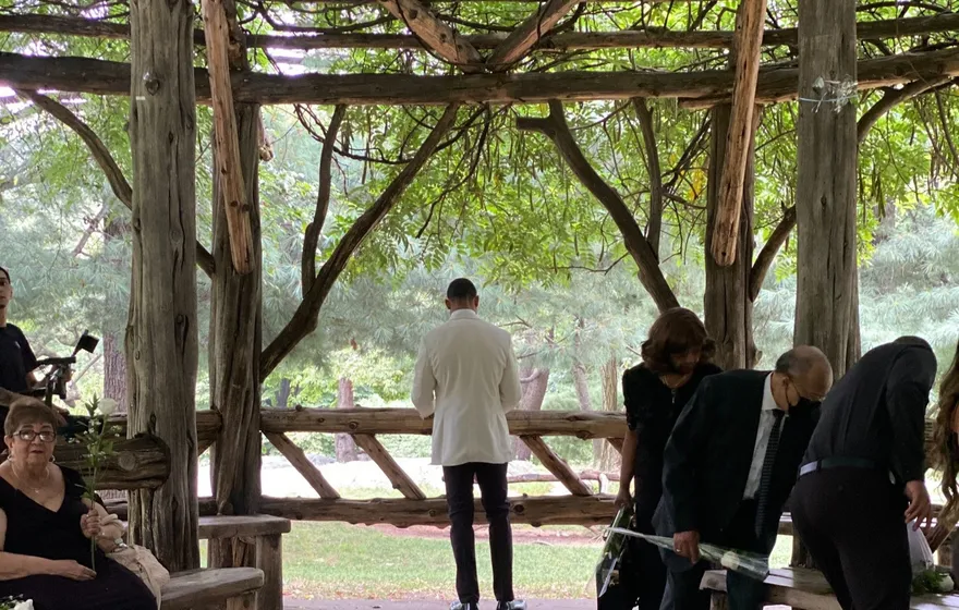 People sit and stand around the cedar benches under the open rustic roof of Cop Cot, a man in a white jacket at the far railing