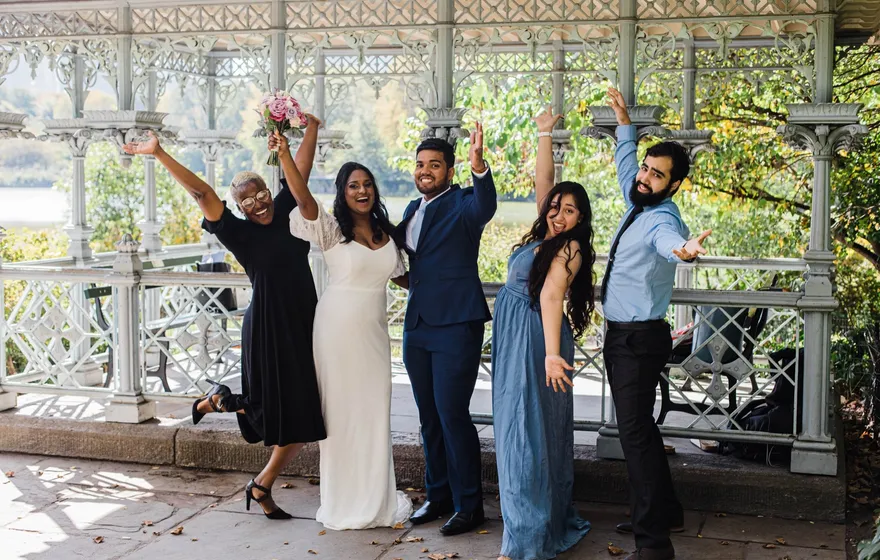 A bride and groom and three others throw their arms up and cheer inside the white iron Ladies Pavilion in Central Park