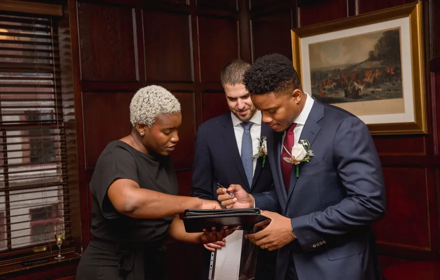 The officiant holds a black folder open while one groom signs and the other leans in to watch, in a wood-paneled room