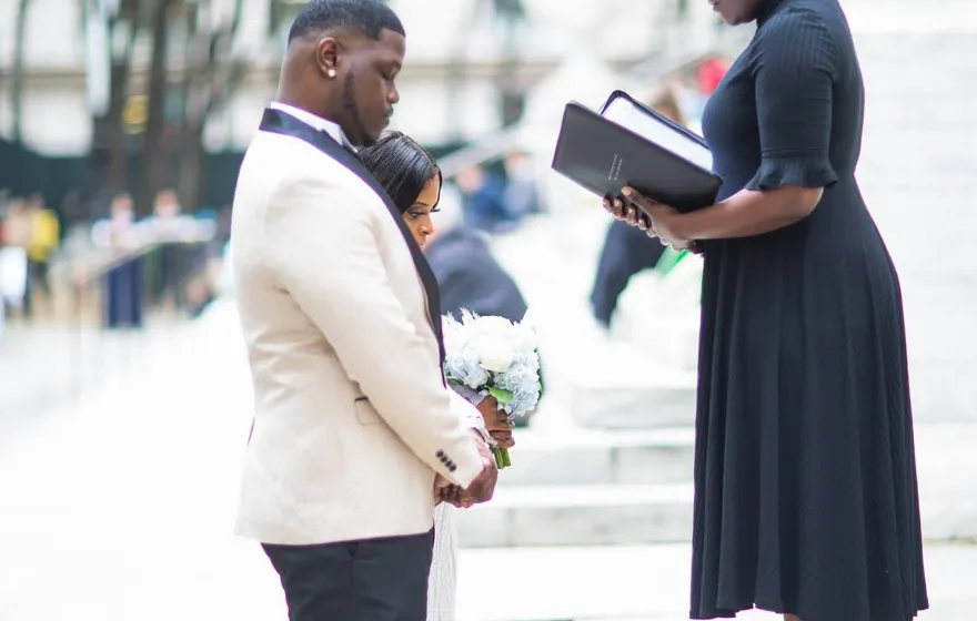On stone steps outside a library, the officiant reads from a black folder while the bride and groom stand close, facing each other