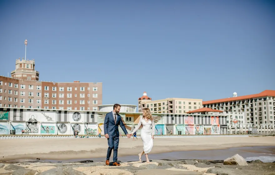 A couple walks hand in hand along the beach with boardwalk buildings behind them