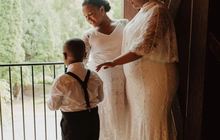 A woman in a lace gown and a girl in a white lace dress stand at an open barn door with a small boy in suspenders