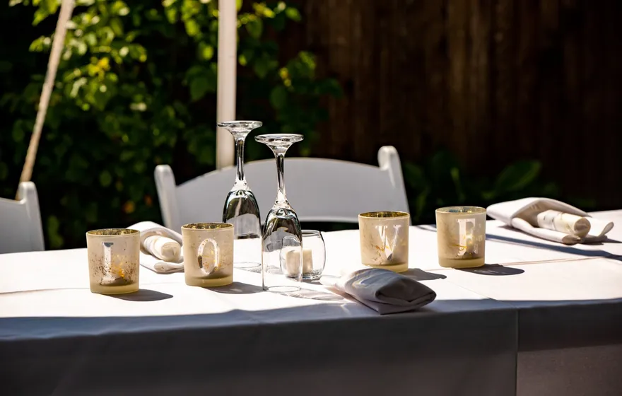 Four glass votive holders spelling LOVE on a white tablecloth at an outdoor wedding, with wine glasses and folded napkins beside them
