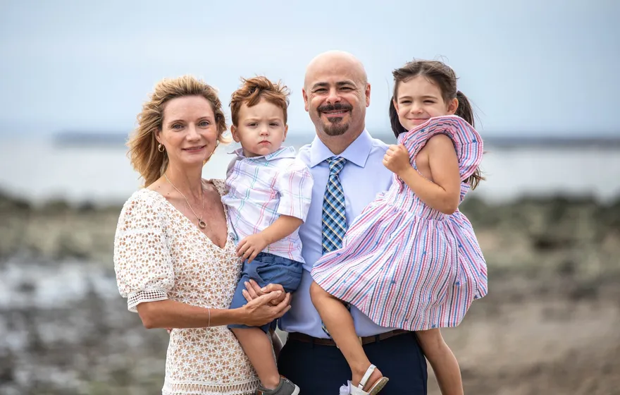 A couple on a rocky beach, the man holding a little girl in a striped dress and the woman holding a toddler in a white shirt