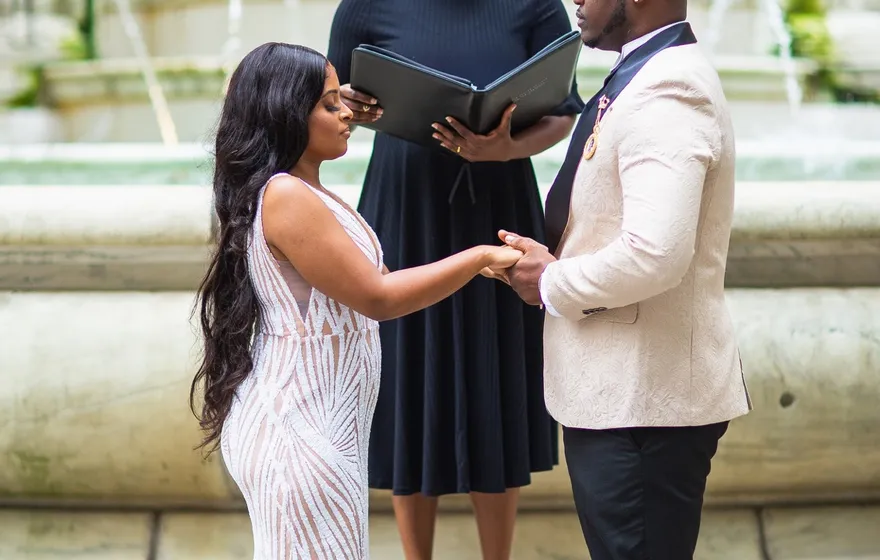 The officiant reads from a black binder at the top of stone steps while the couple holds hands in front of her, a fountain behind them