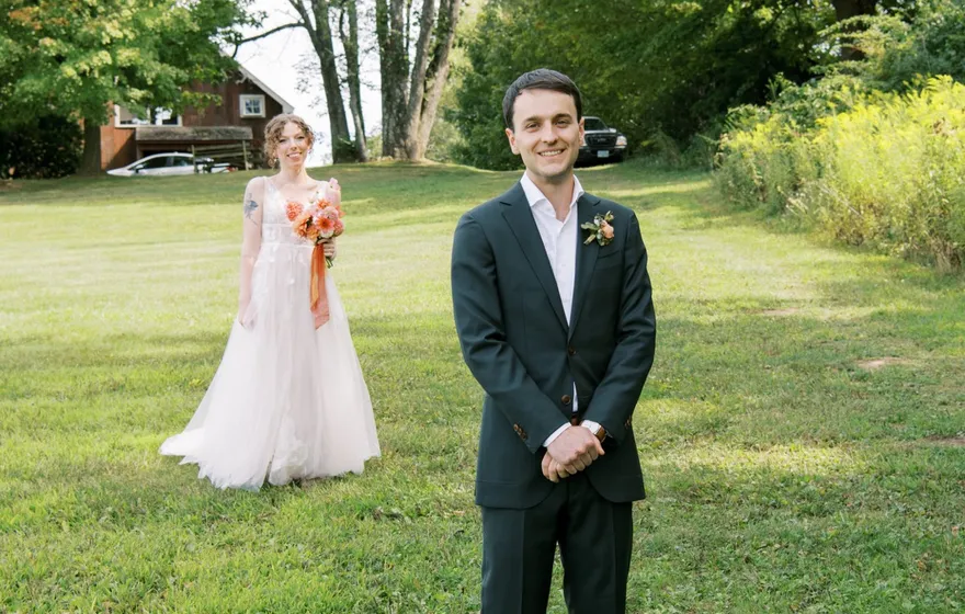 A groom in a dark suit stands in a field with his back to the bride, who walks toward him across the grass in her gown