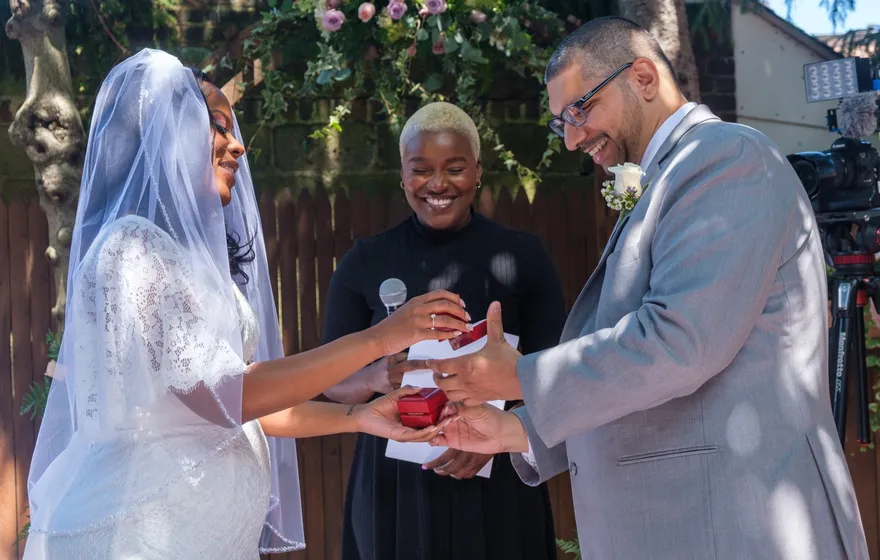 A bride in a lace dress and veil and a groom in a gray suit hold out small red ring boxes to each other while the officiant smiles between them