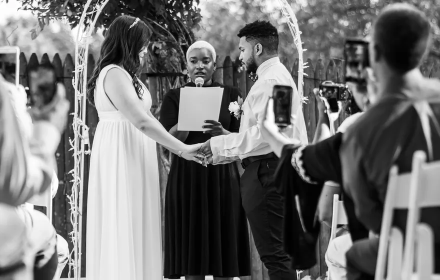 Black and white photo of a backyard ceremony seen down a petal-strewn aisle: the officiant holds her script while the couple holds hands under a wire arch and guests raise their phones