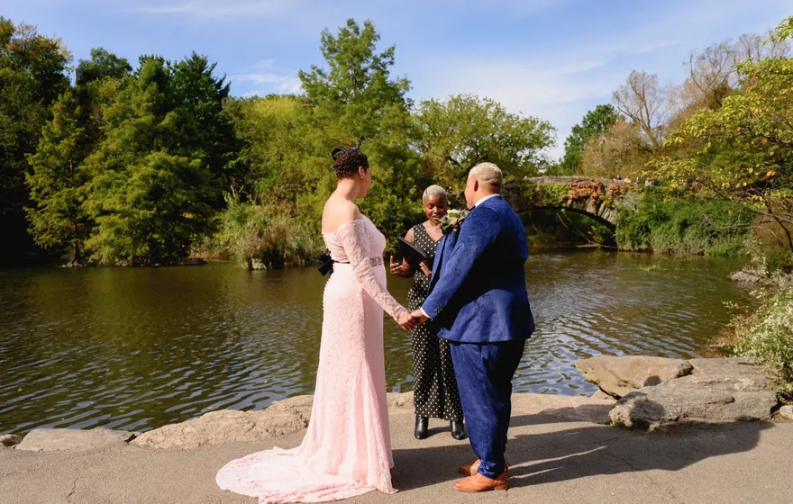 A couple in a pink lace gown and a blue suit hold hands on the rocks by the pond in Central Park while the officiant stands between them, Gapstow Bridge behind