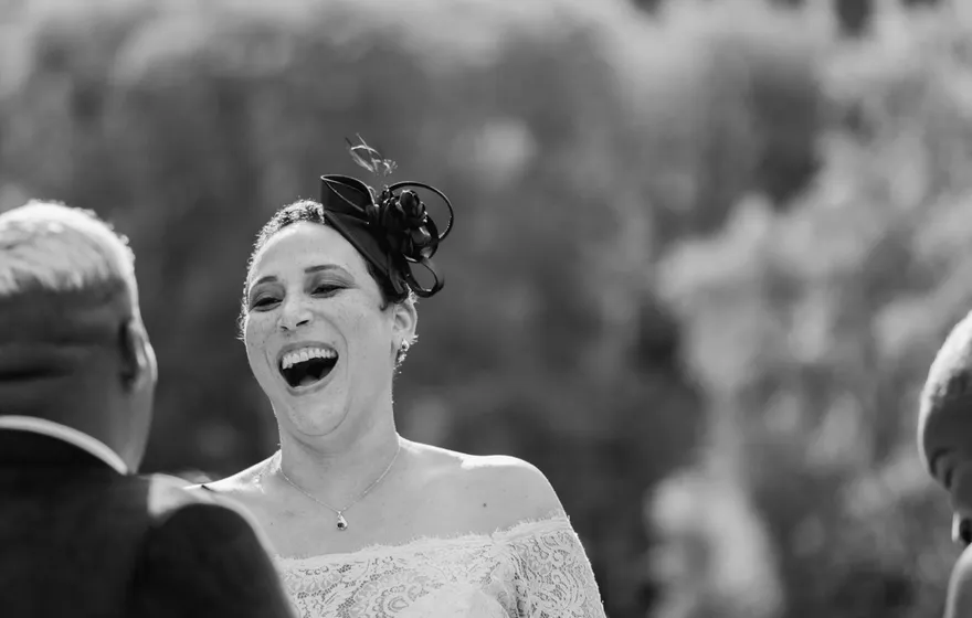Black and white photo of a laughing bride in a lace gown and fascinator at a Central Park vow renewal, the officiant's open binder in the foreground and skyscrapers behind