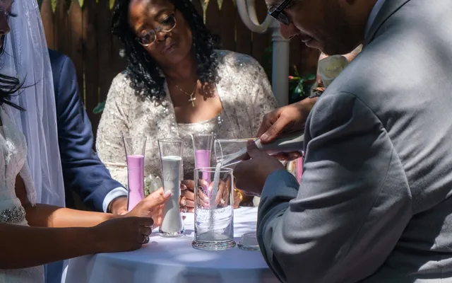 A woman pours sand into a shared glass vessel beside the groom at a small table set with sand vases