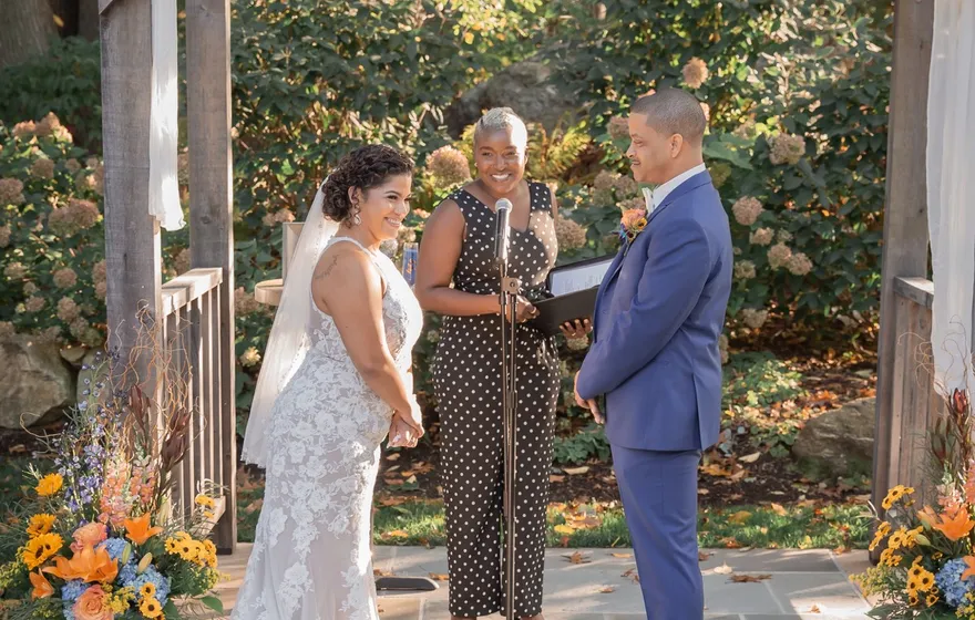 A bride and groom face each other under a white-draped garden pergola while the officiant stands between them reading from a folder