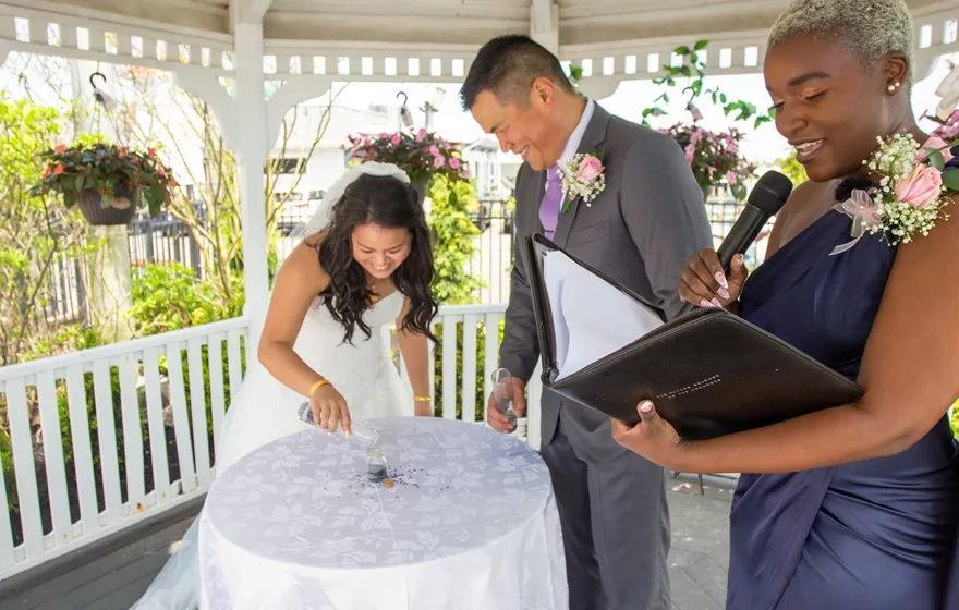 In a white gazebo, a bride leans over a small round table with a white cloth while the officiant speaks into a microphone beside her and the groom holds a folder