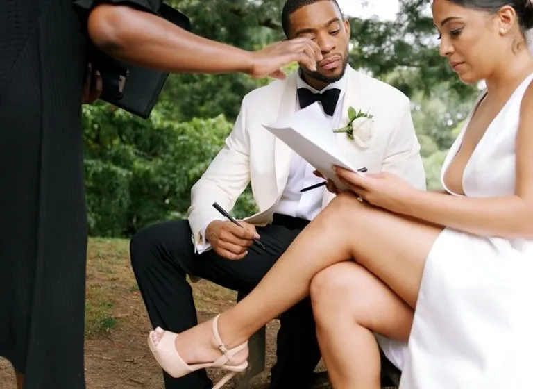 On a park bench, the officiant leans over to point out a line on the marriage license while the groom signs it on his knee and the bride sits beside him