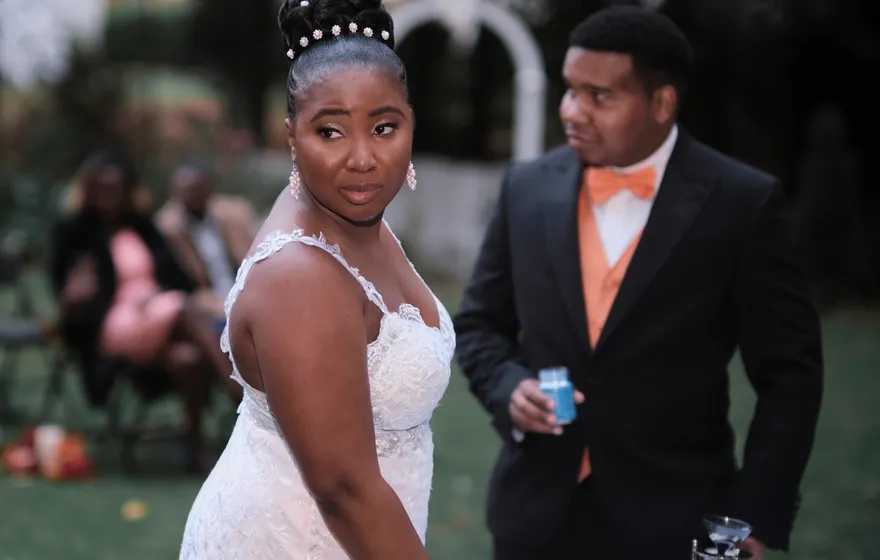 A groom in a black suit holds a small jar of blue sand while his bride looks back over her shoulder