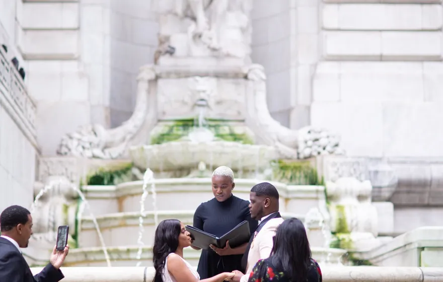 A couple stands before the officiant at the fountain on the steps of the New York Public Library while one guest films and another looks on