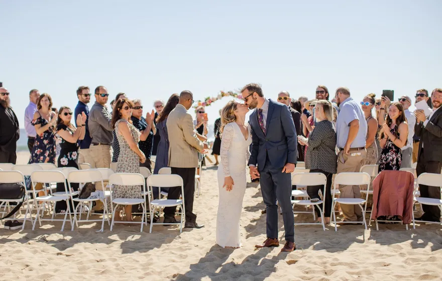 A bride and groom kiss in the aisle on a beach while guests stand and applaud behind rows of white chairs