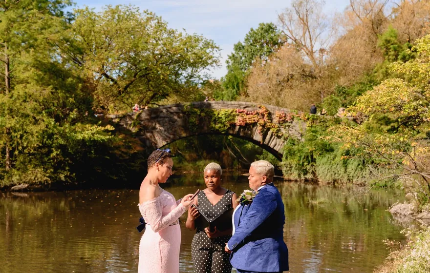 A wide view of a small ceremony at the edge of the Pond, the officiant between two partners in a pink gown and a blue suit, with Gapstow Bridge in the trees to the left